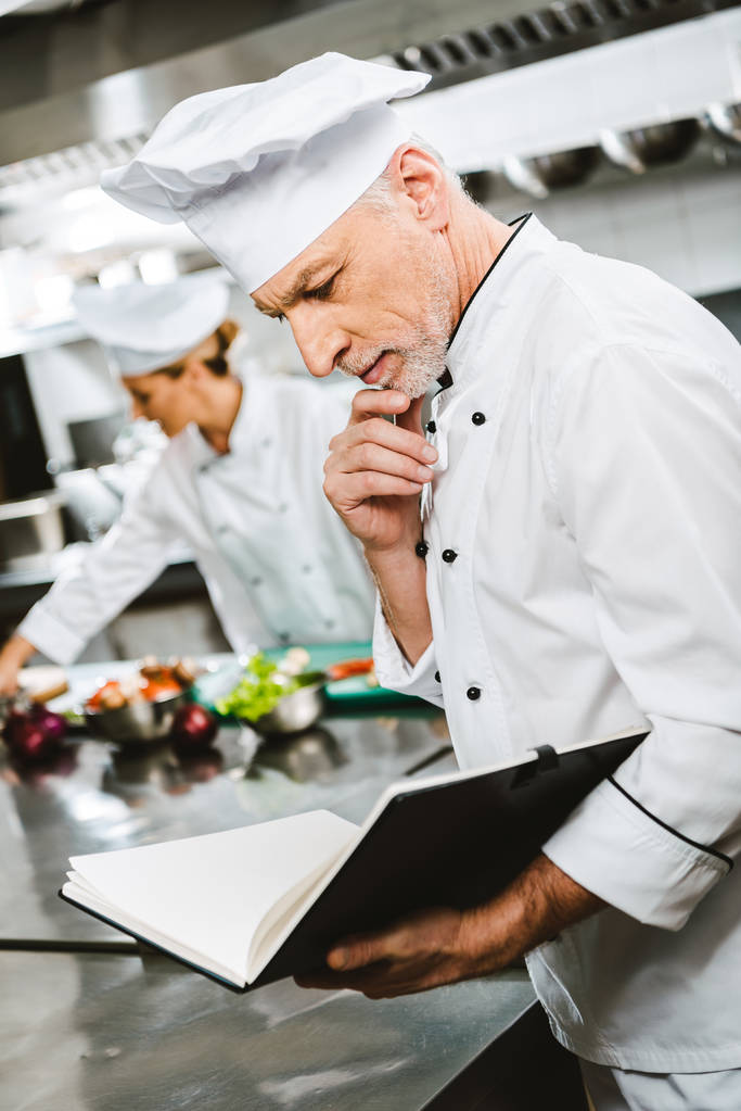 красивый вдумчивый мужчина шеф-повар поддерживает подбородок рукой, читая книгу рецептов на кухне ресторана
 - Фото, изображение
