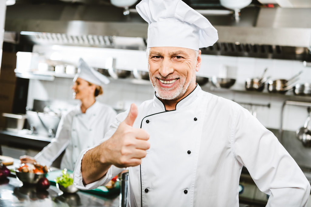 επιλεκτική εστίαση του όμορφος χαμογελώντας αρσενικό σεφ σε ομοιόμορφη κοιτάζοντας κάμερα και δείχνει τον αντίχειρα επάνω σημάδι σε κουζίνα εστιατορίου - Φωτογραφία, εικόνα