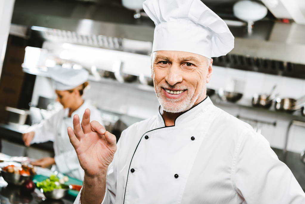szelektív összpontosít, szép mosolygó férfi szakács egységes látszó-on fényképezőgép, és bemutatja az ok jel az étterem konyha - Fotó, kép