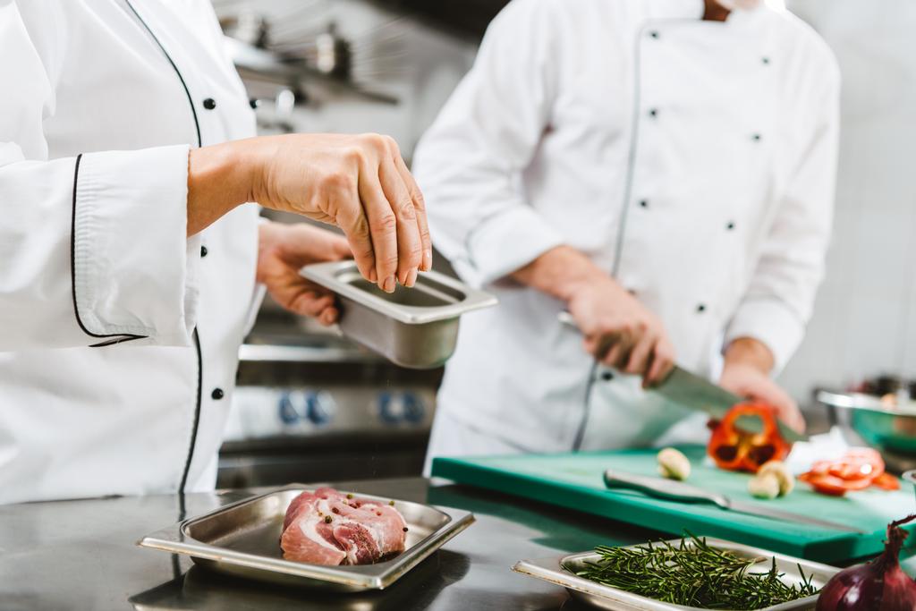 περικοπεί δείτε θηλυκό σεφ καρυκεύματα κρέας ενώ ο άνθρωπος μαγείρεμα σε φόντο σε κουζίνα εστιατορίου - Φωτογραφία, εικόνα