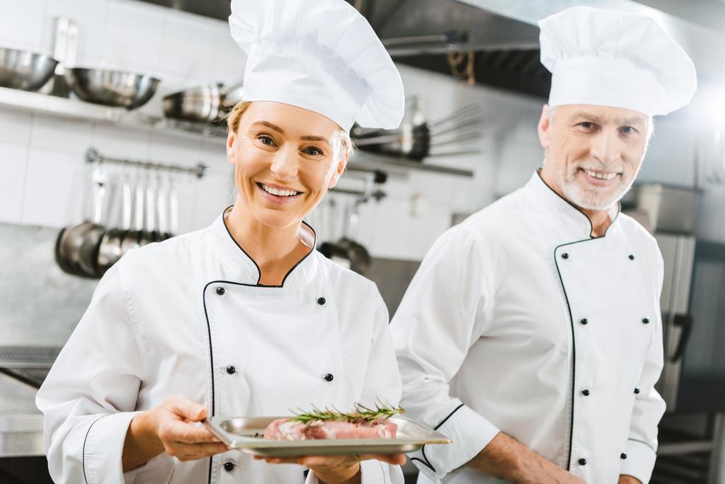 female and male chefs in uniform with meat steak on plate looking at camera in restaurant kitchen - Photo, Image