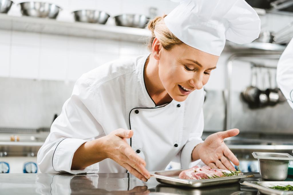 hermosa hembra sonriente chef en uniforme sosteniendo plato con plato de carne en la cocina del restaurante
 - Foto, imagen