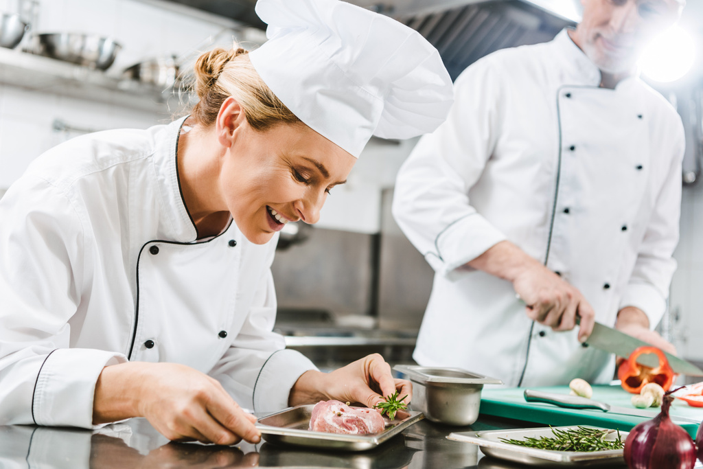 kadın şef meslektaşım üniformalı arka planda lokanta mutfağı pişirme sırasında et çanak tutan gülümseyerek - Fotoğraf, Görsel