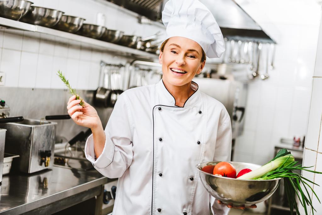 schöne lächelnde Köchin in Uniform mit Rosmarin und Schüssel mit Gemüse in der Restaurantküche - Foto, Bild