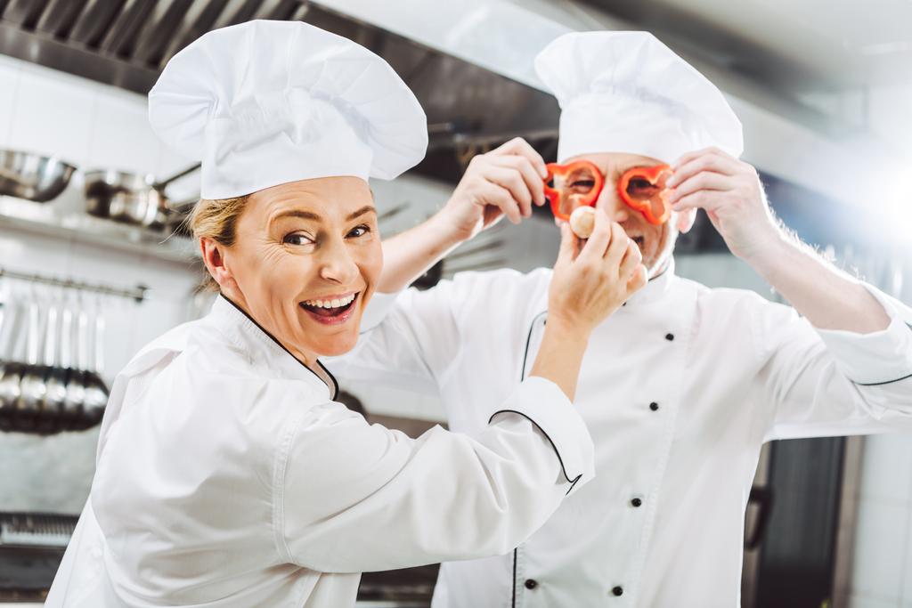 vrouwelijke kok kijken camera terwijl mannelijke chef-kok peper segmenten voor gezicht houden in restaurant keuken - Foto, afbeelding