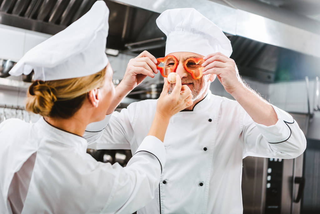 жінка готує, дивлячись на чоловічий шеф-кухар, що тримає шматочки перцю перед обличчям на кухні ресторану
 - Фото, зображення