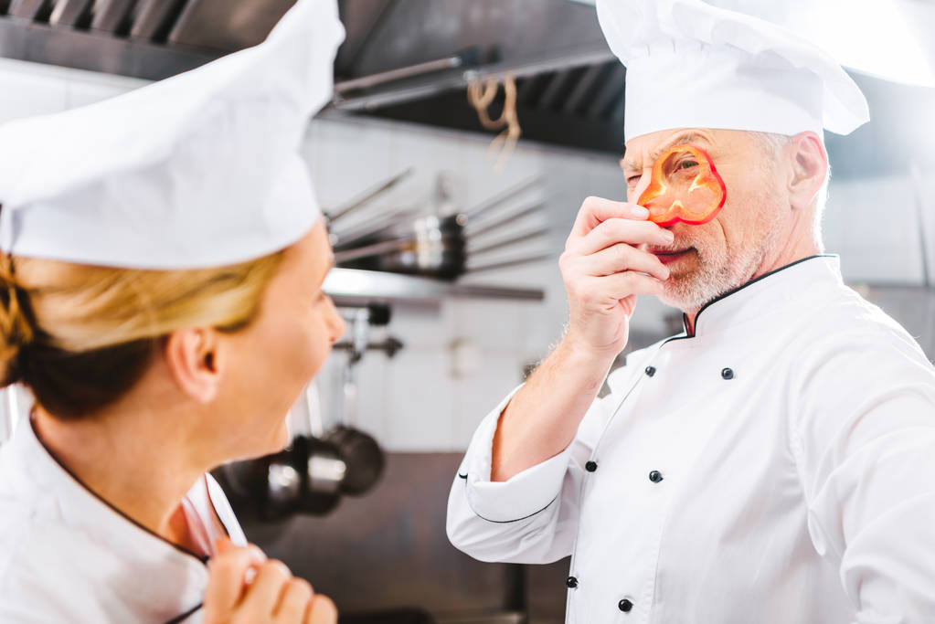 cocinero femenino mirando al chef masculino sosteniendo rebanada de pimienta frente a la cara en la cocina del restaurante
 - Foto, Imagen