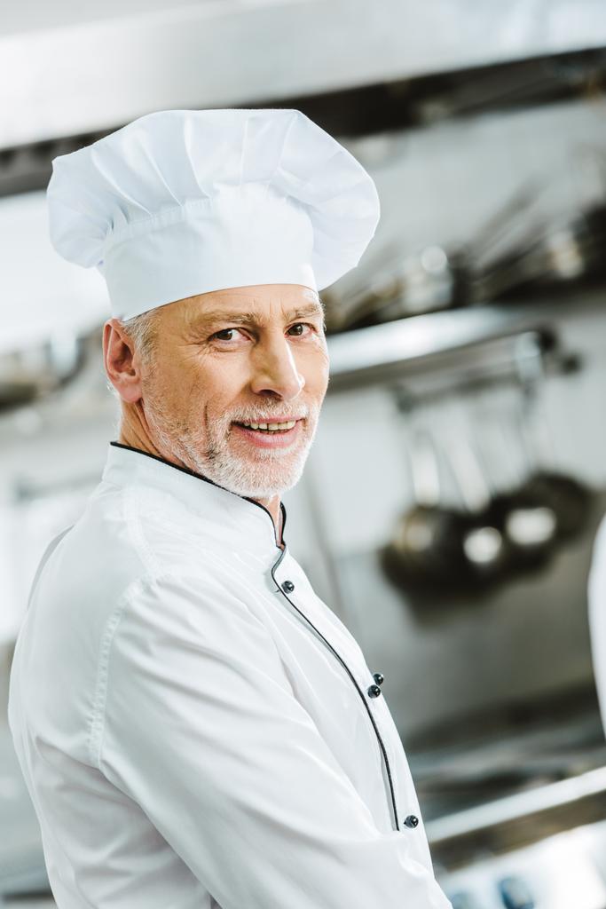 όμορφος χαμογελώντας αρσενικό σεφ με τη στολή και καπάκι βλέπουν φωτογραφική μηχανή σε κουζίνα εστιατορίου - Φωτογραφία, εικόνα