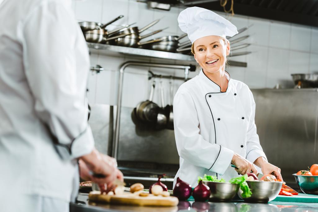 kaunis naiskokki univormussa hymyilee ruoanlaitossa ravintolan keittiössä
 - Valokuva, kuva