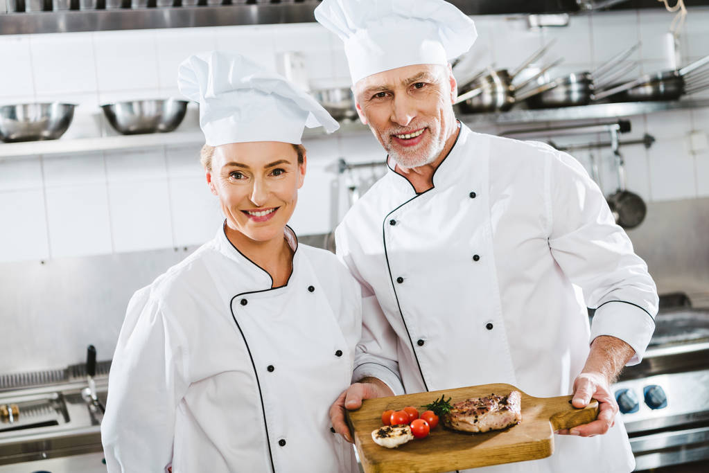 жіночі та чоловічі шеф-кухарі в уніформі, дивлячись на камеру, представляючи м'ясну тарілку на дерев'яній дошці на кухні ресторану
 - Фото, зображення