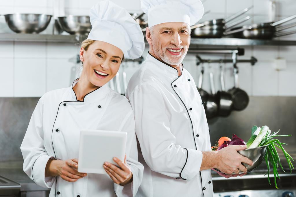 жіночі та чоловічі шеф-кухарі у формі проступків, дивлячись на камеру та використовуючи цифровий планшет під час приготування їжі на кухні ресторану
 - Фото, зображення