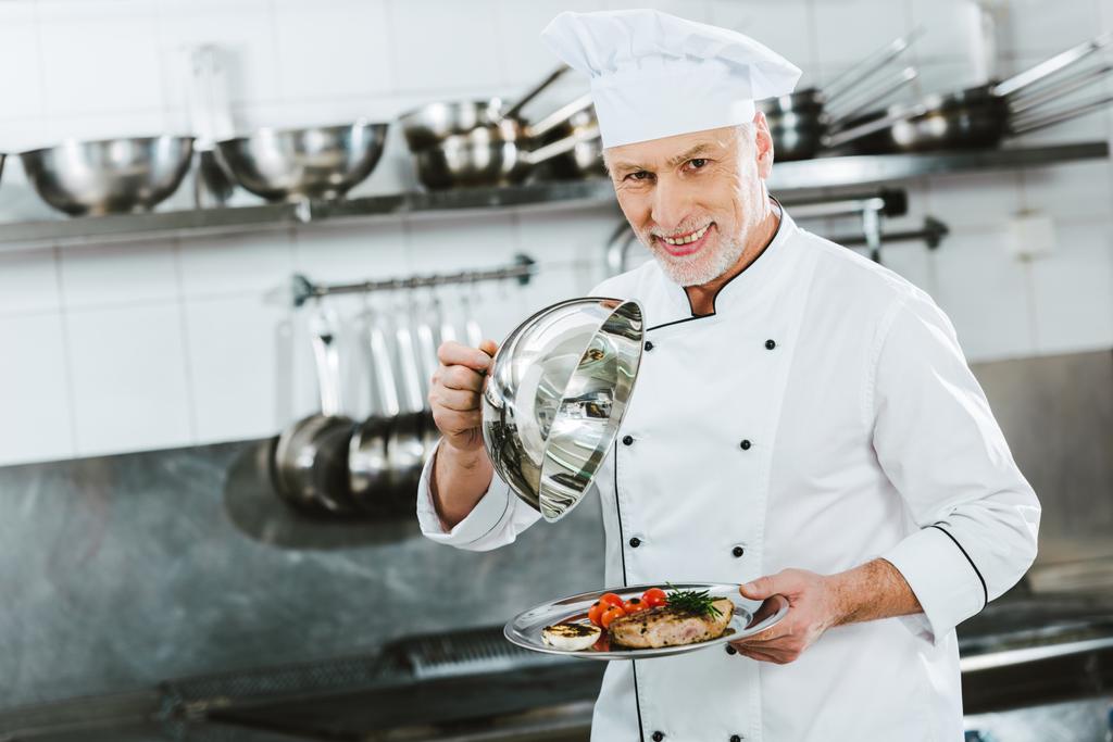 schöner männlicher Koch in Uniform schaut in die Kamera und hält Kuppel vom Serviertablett mit Fleischplatte in der Restaurantküche - Foto, Bild