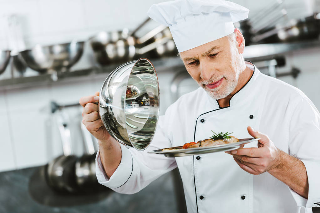 selektiver Fokus eines gut aussehenden männlichen Kochs in Uniform, der in der Restaurantküche Tablett mit Fleischplatte hält - Foto, Bild