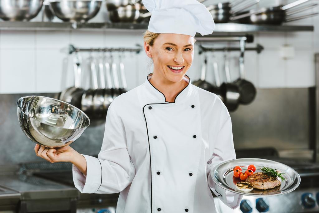 beautiful smiling female chef in uniform holding dome from serving tray with meat dish in restaurant kitchen - Photo, Image