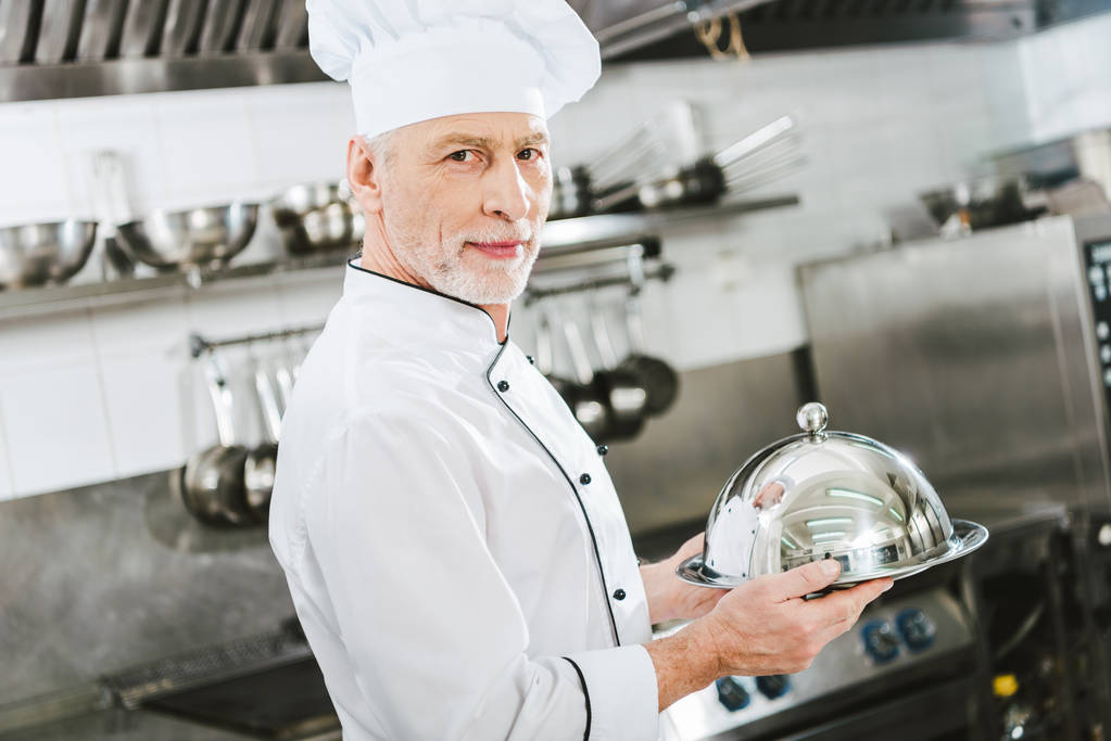 όμορφος αρσενικό σεφ σε ομοιόμορφο κοιτάζοντας κάμερα και κρατώντας το δίσκο σερβιρίσματος με θόλο σε κουζίνα εστιατορίου - Φωτογραφία, εικόνα