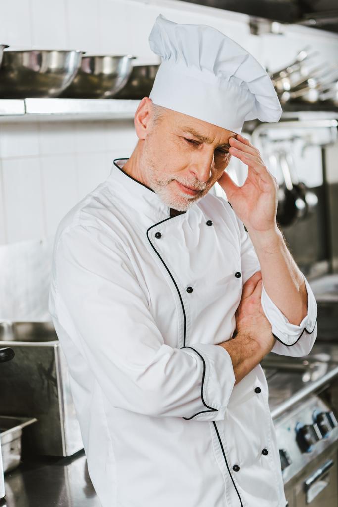 Müder männlicher Koch in Uniform berührt Kopf und hat Kopfschmerzen in Restaurantküche - Foto, Bild
