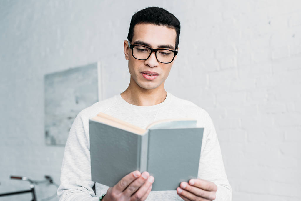 young mixed race serious man in white sweatshirt and glasses reading book - Photo, Image