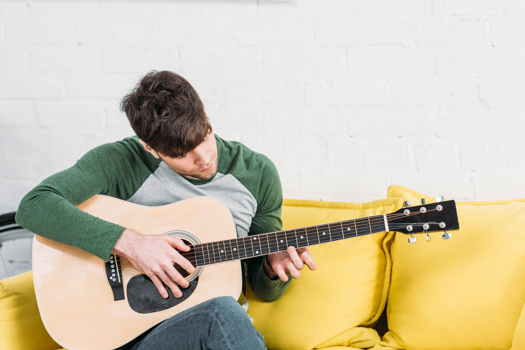 młody człowiek siedzi na kanapie żółty i gra gitara akustyczna - Zdjęcie, obraz
