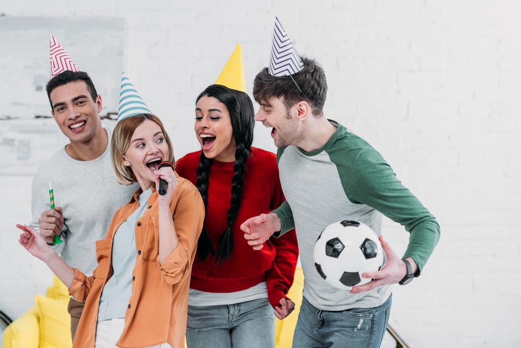 fröhliche multiethnische Freunde in bunten Papierhüten, die zu Hause Party machen und Karaoke singen - Foto, Bild