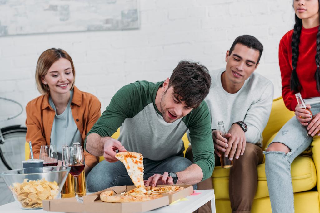 junger Mann nimmt ein Stück Pizza, während multikulturelle Freunde Drinks genießen - Foto, Bild