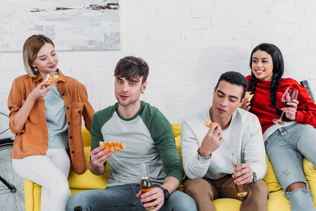 onnelliset monikulttuuriset ystävät syövät pizzaa ja nauttivat juomia istuessaan yhdessä keltaisella sohvalla
 - Valokuva, kuva