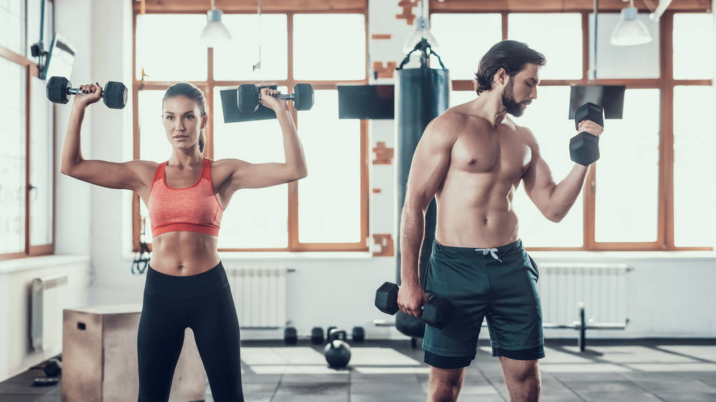Girl And Guy In Gym Doing Dumbbells Exercises. Training Day. Fitness Club. Healthy Lifestyle. Powerful Athletes. Active Holidays. Crossfit Concept. Sportsman Without T Shirt. Sunny Day. - Photo, Image