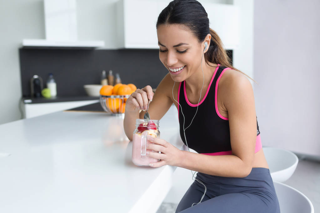Tiro de jovem esportivo comendo smoothie de morango enquanto ouve música na cozinha em casa
. - Foto, Imagem