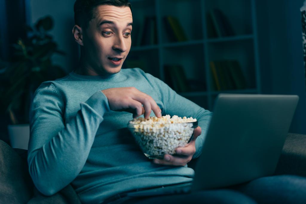 překvapený muž sledování filmu na notebooku a doma drží misku popcornu - Fotografie, Obrázek