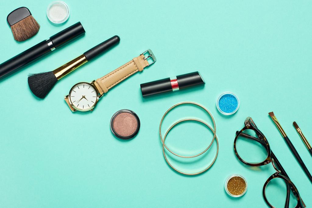 Vista superior de rímel, relógio, batom, pulseiras, sombra, blush, óculos e escovas cosméticas em fundo turquesa
 - Foto, Imagem