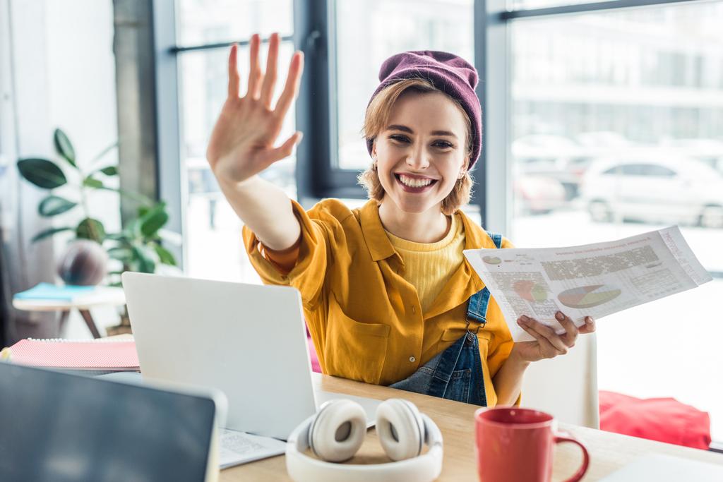 Lächelnde junge Fachfrau, die in die Kamera blickt, winkt und Dokumente in der Hand hält, während sie am Computertisch im Loftbüro sitzt  - Foto, Bild