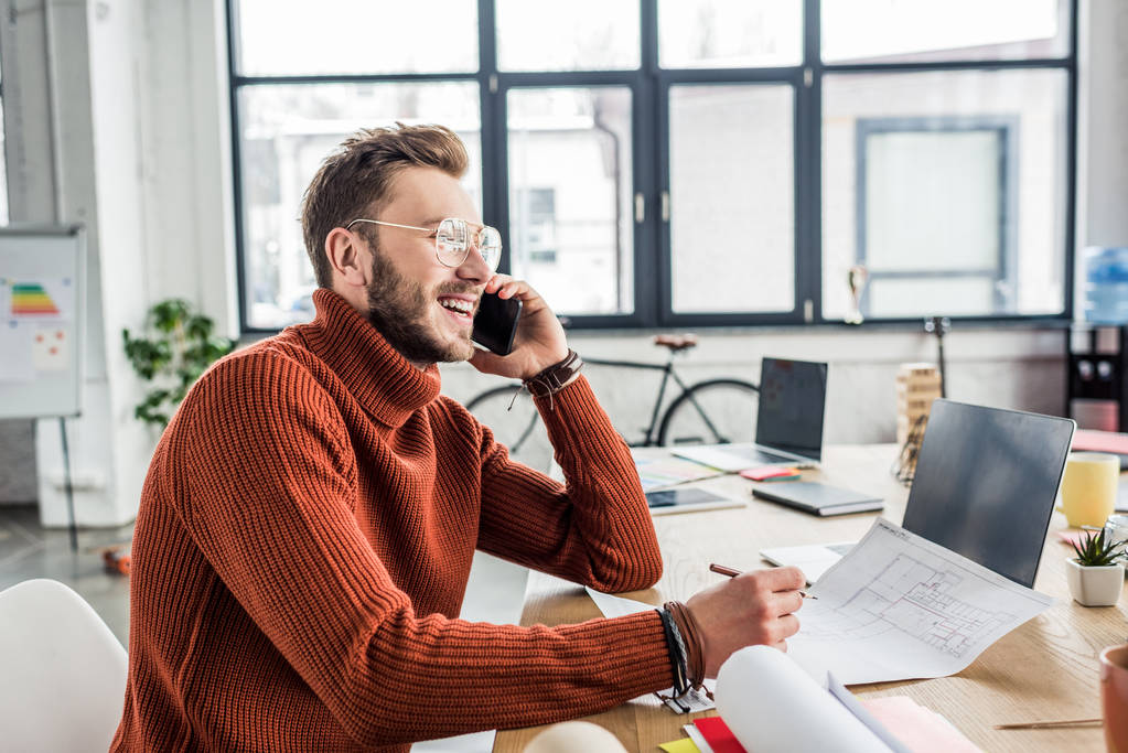 улыбающийся мужчина-архитектор, сидящий за столом, разговаривающий на смартфоне и работающий над чертежами в лофт-офисе
 - Фото, изображение