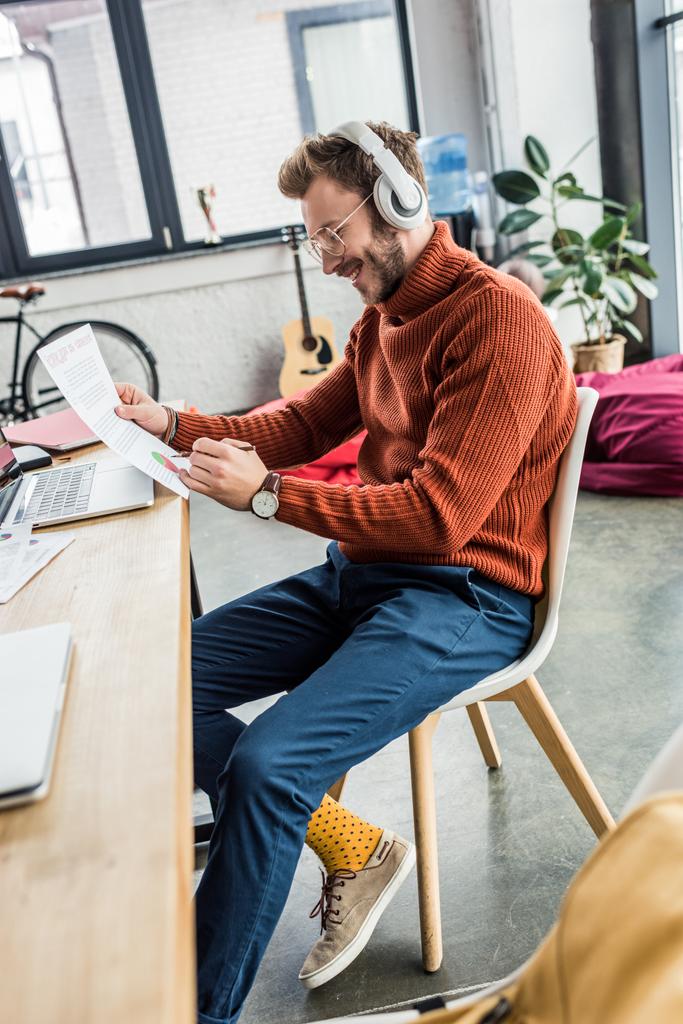guapo hombre de negocios casual sentado en el escritorio en los auriculares y trabajando en el documento en la oficina loft
 - Foto, Imagen