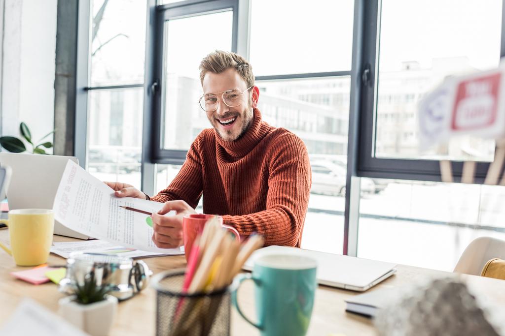 улыбающийся бизнесмен в очках, сидящий за столом и работающий над документом в лофт-офисе
 - Фото, изображение