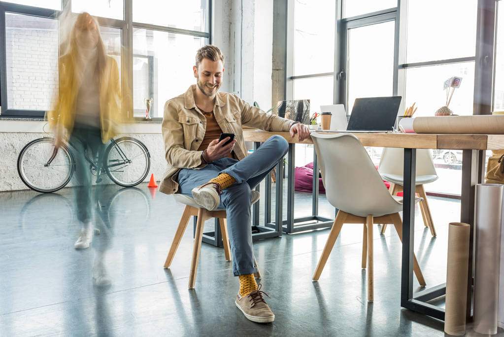 männlicher Architekt sitzt und Smartphone mit Kollegen in Bewegung Unschärfe auf Hintergrund in Loft-Büro - Foto, Bild
