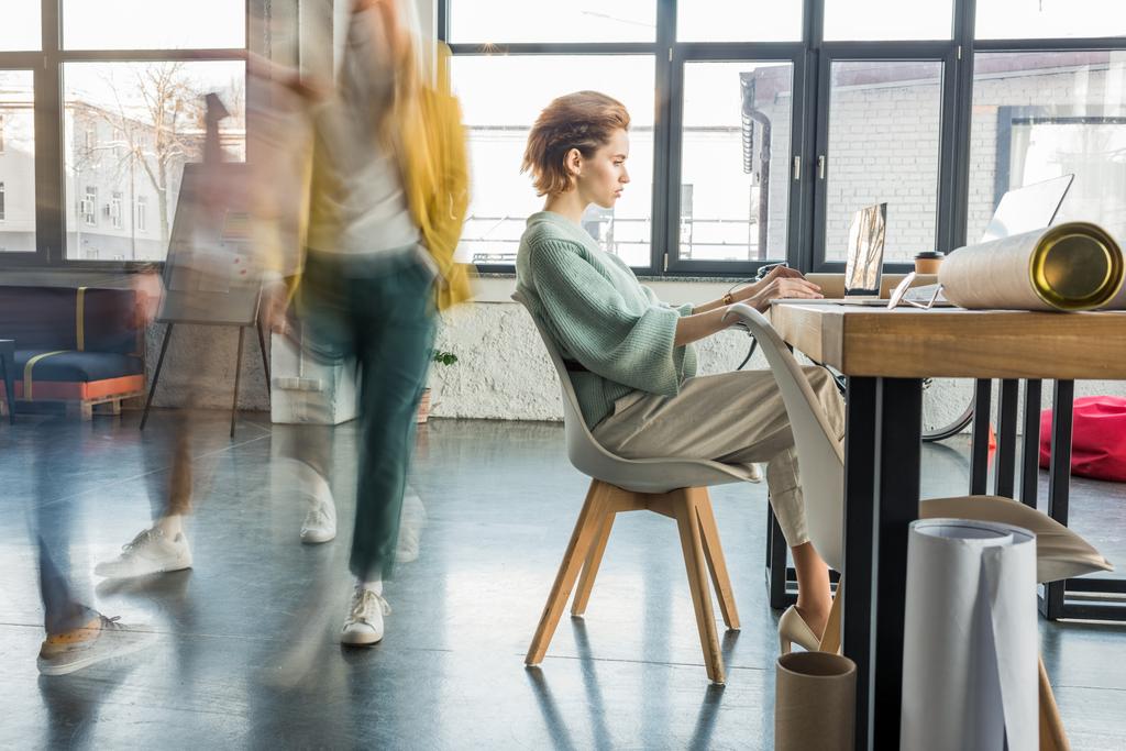 背景にぼかしの動きで同僚とロフト オフィスのデスクでノート パソコンを使用して座っている女性建築家  - 写真・画像