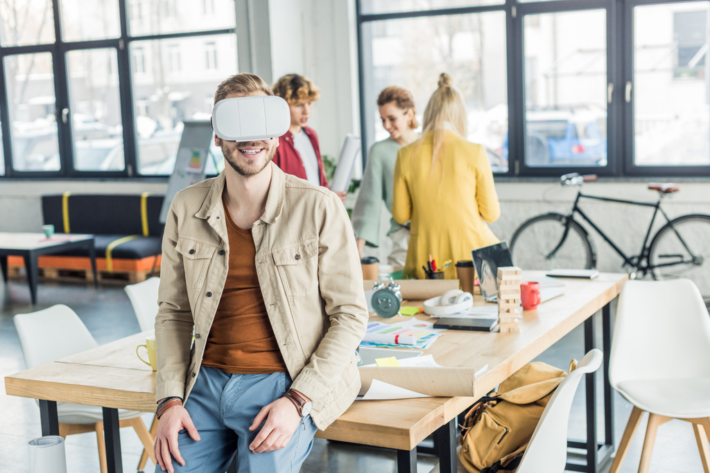 lächelnder männlicher Designer mit Virtual-Reality-Erfahrung im Loft-Büro mit Kollegen im Hintergrund - Foto, Bild