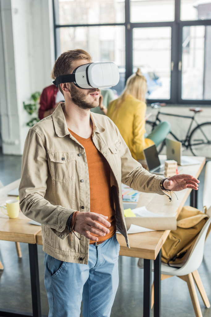 male designer gesturing with hands while having virtual reality experience in loft office with colleagues on background - Photo, Image