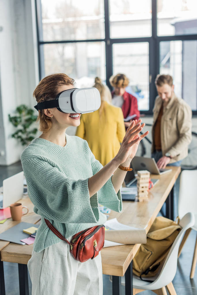 sorridente designer feminino gestos com as mãos, tendo experiência realidade virtual no escritório loft com colegas em segundo plano
 - Foto, Imagem