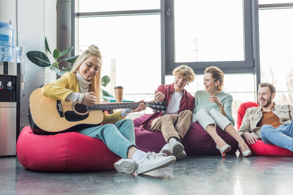 fröhliche Gruppe von Freunden auf Sitzsäcken sitzend und Gitarre spielend  - Foto, Bild