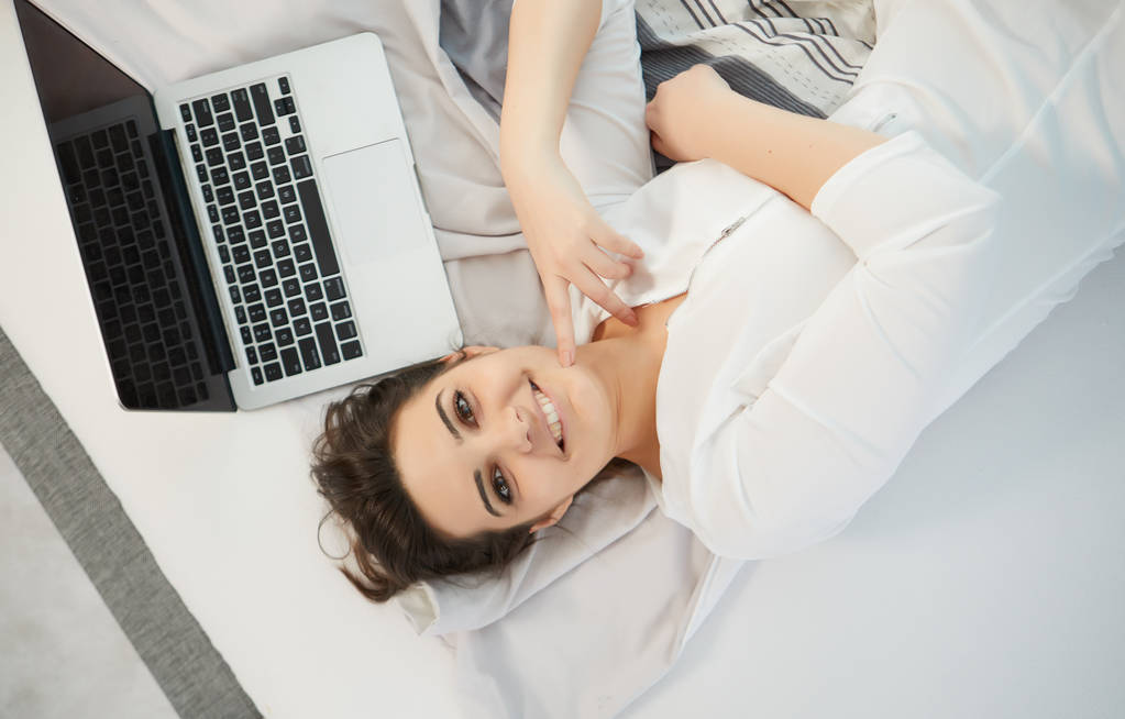 Молодая счастливая женщина отдыхает в своей кровати, она подключается к ноутбуку, вид сверху
  - Фото, изображение