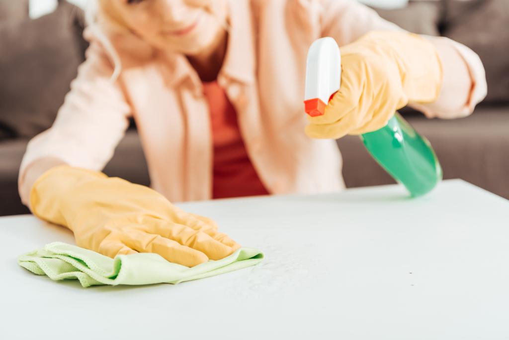 Vista parcial de la mujer en guantes de goma amarilla superficie de la mesa de limpieza con trapo y spray
  - Foto, Imagen