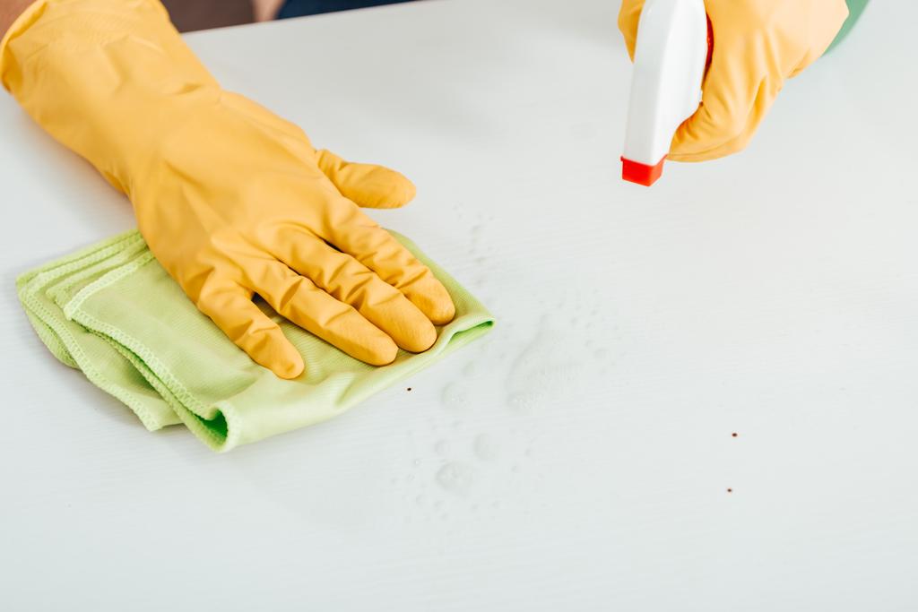 Vista recortada de la mujer en guantes de goma amarilla limpiando la superficie blanca con trapo y spray
 - Foto, imagen