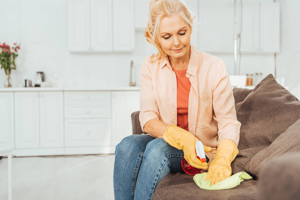 Mujer mayor en guantes de goma limpiando sofá con spray y trapo
 - Foto, Imagen