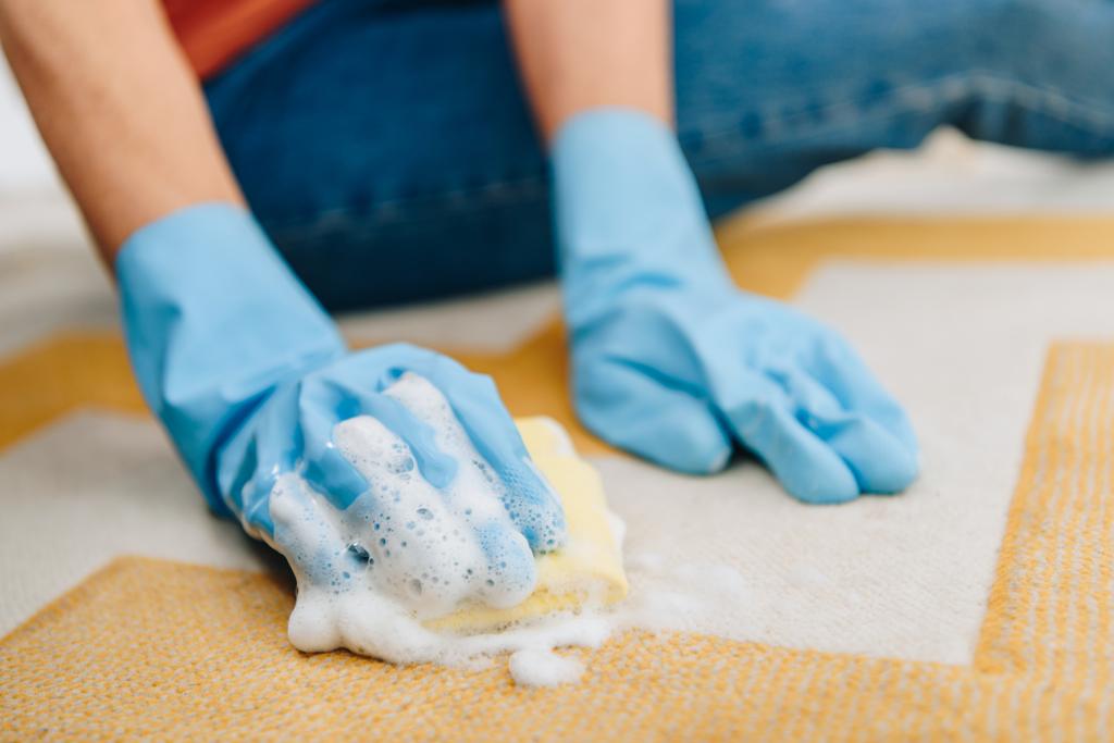 Gedeeltelijke weergave van vrouw in rubberen handschoenen schoonmaak gestreepte tapijt met schuimend spons - Foto, afbeelding