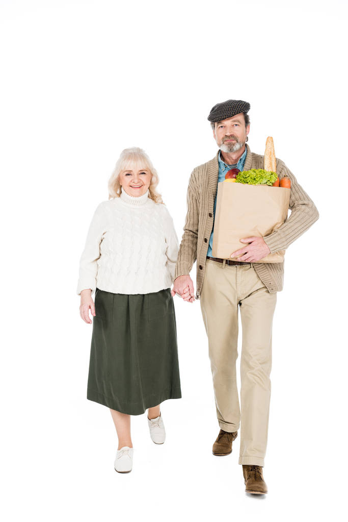 cheerful senior man holding hands with wife while holding paper bag with products isolated on white - Photo, Image