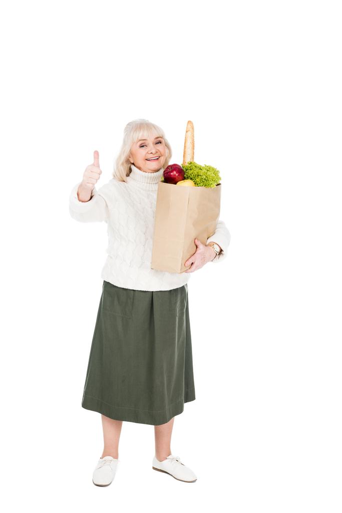 Veselý senior žena držící papírový sáček s potravinami a ukazuje palcem nahoru izolované na bílém - Fotografie, Obrázek
