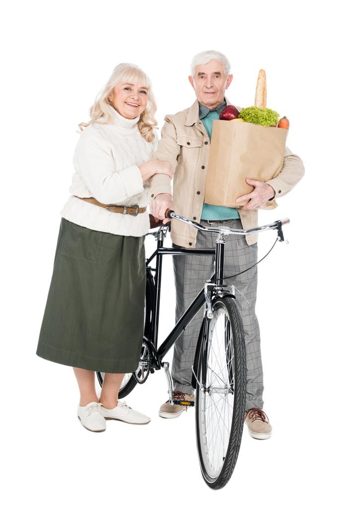 feliz esposa de pie con el marido jubilado sosteniendo bicicleta y bolsa de papel aislado en blanco
 - Foto, Imagen