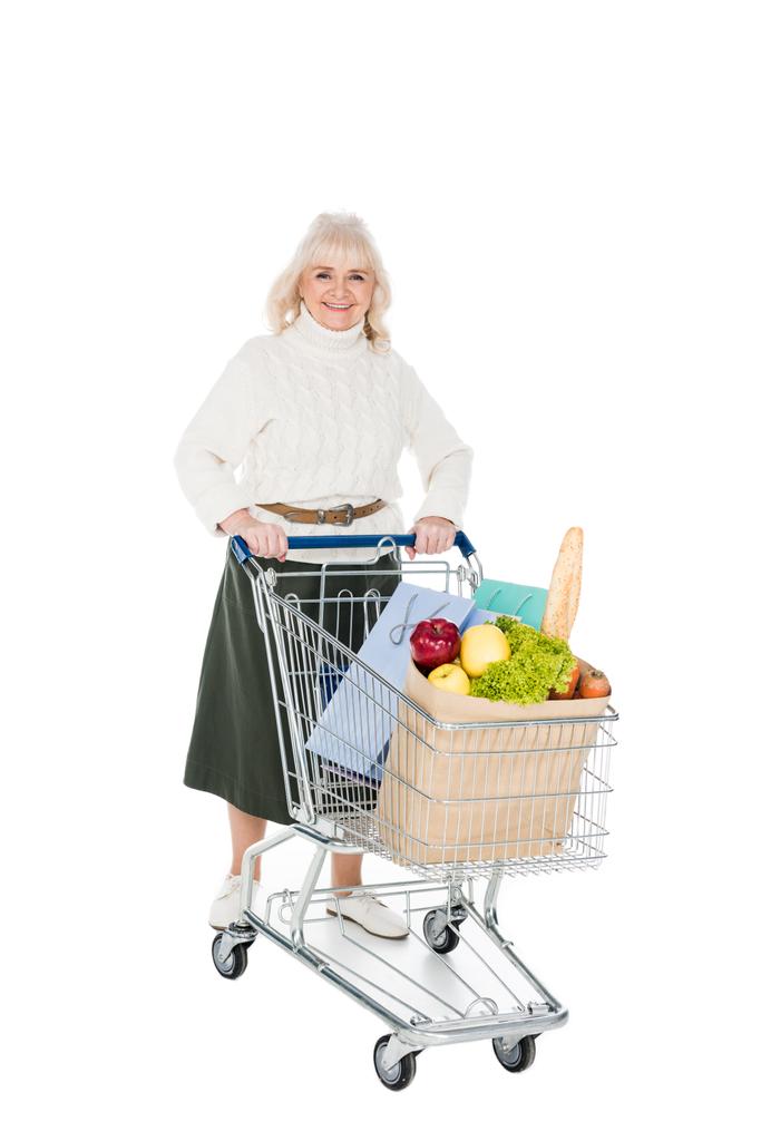 mulher sênior feliz segurando carrinho de compras com sacos de compras e saco de papel com mantimentos isolados em branco
 - Foto, Imagem