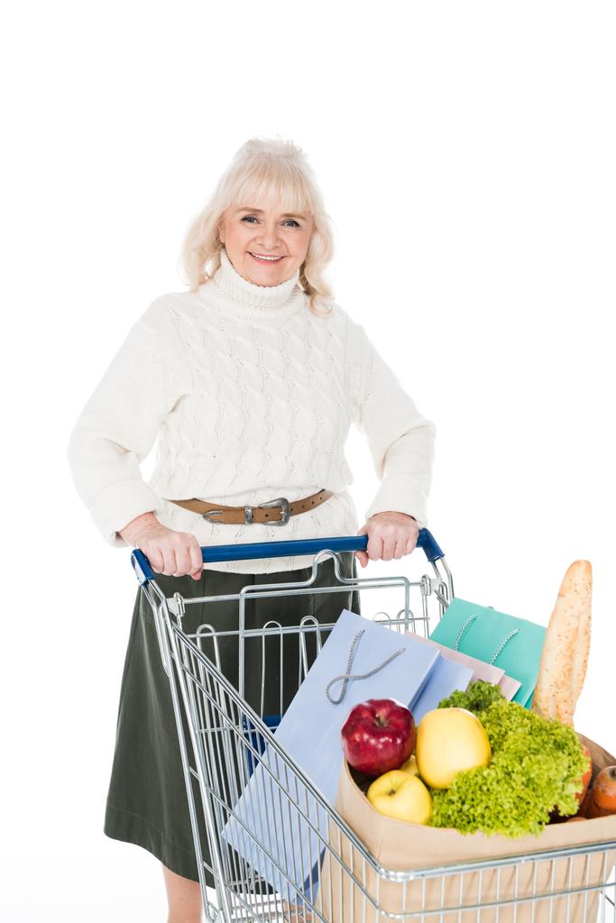 sonriente mujer mayor sosteniendo carrito de compras con bolsas de compras y bolsa de papel con comestibles aislados en blanco
 - Foto, imagen
