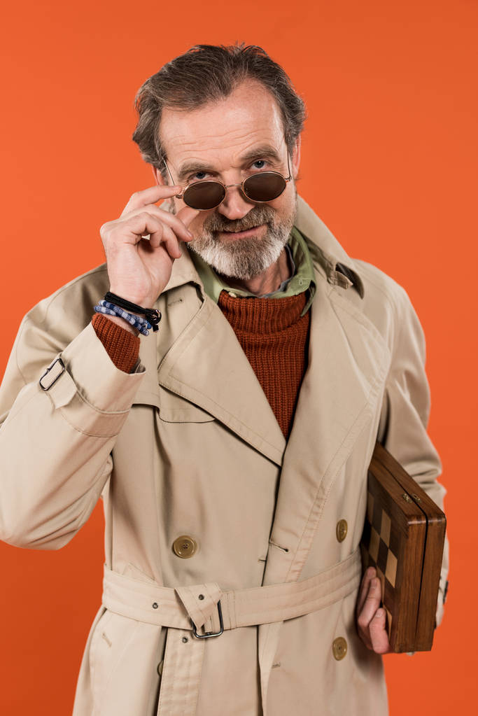 модный пожилой человек трогает солнцезащитные очки, держа шахматную доску изолированной на оранжевом
  - Фото, изображение